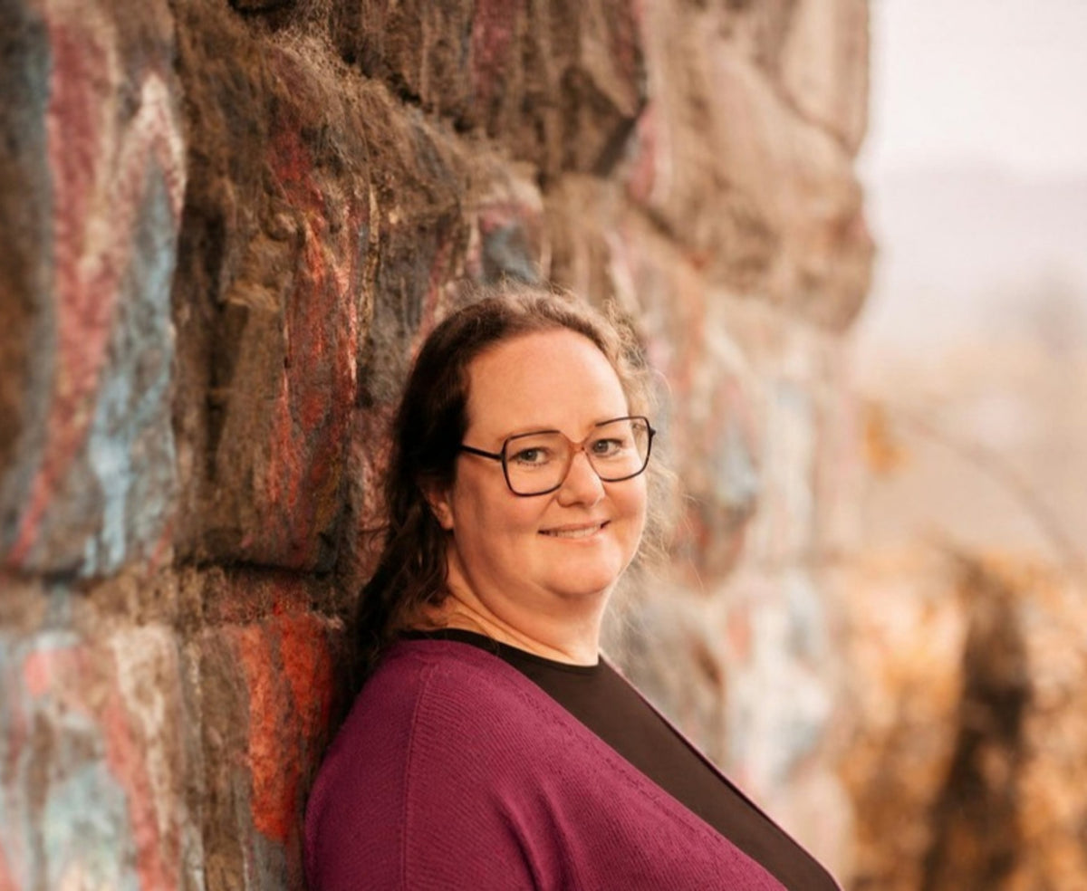 Nicola Forth lächelnd vor einer bemalten Steinwand, in Jacke in Brombeere und Oberteil in Espresso